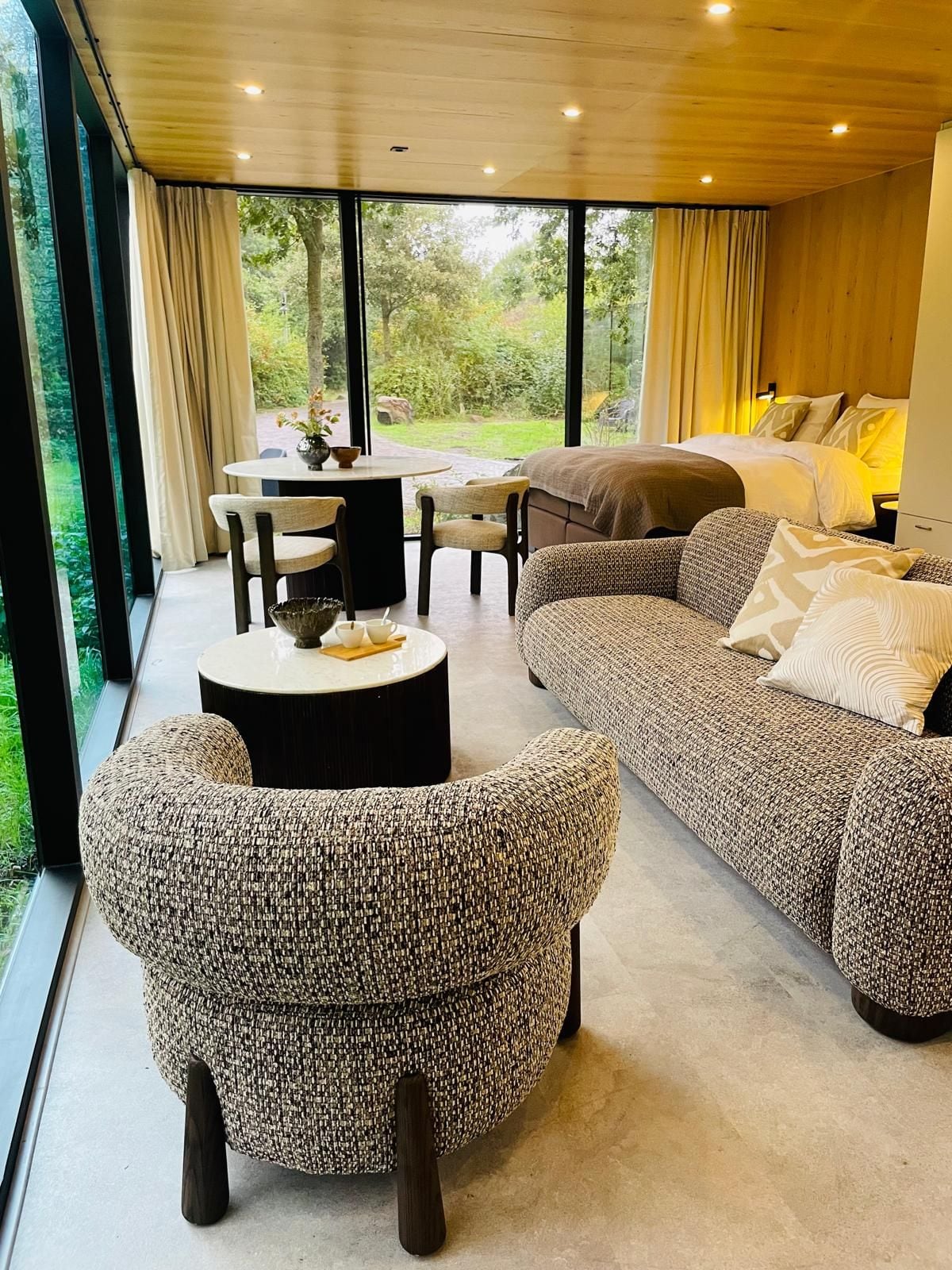 Uma sala de estar moderna com um sofá grande, uma mesa de centro redonda e uma poltrona de vime. Há uma área de jantar com mesa e cadeiras, e vista para o verde lá fora.