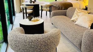 Un salon moderne avec un grand canapé, une table basse ronde et un fauteuil en osier. On y trouve également un coin repas avec une table et des chaises, et une vue sur la verdure à l’extérieur.