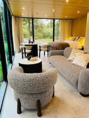 A modern living room with a large sofa, a round coffee table, and a wicker armchair. There is a dining area with a table and chairs, and a view of greenery outside. - S.P.A. Parel in 't Groen (Moergestel)
