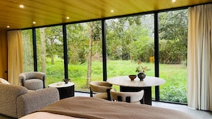 Une chambre avec un grand lit, une table ronde et deux chaises, donnant sur un jardin luxuriant grâce à de grandes fenêtres.