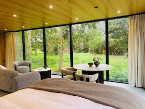 A bedroom with a large bed, a round table, and two chairs, with a view of a lush garden through large windows. - S.P.A. Parel in 't Groen (Moergestel)
