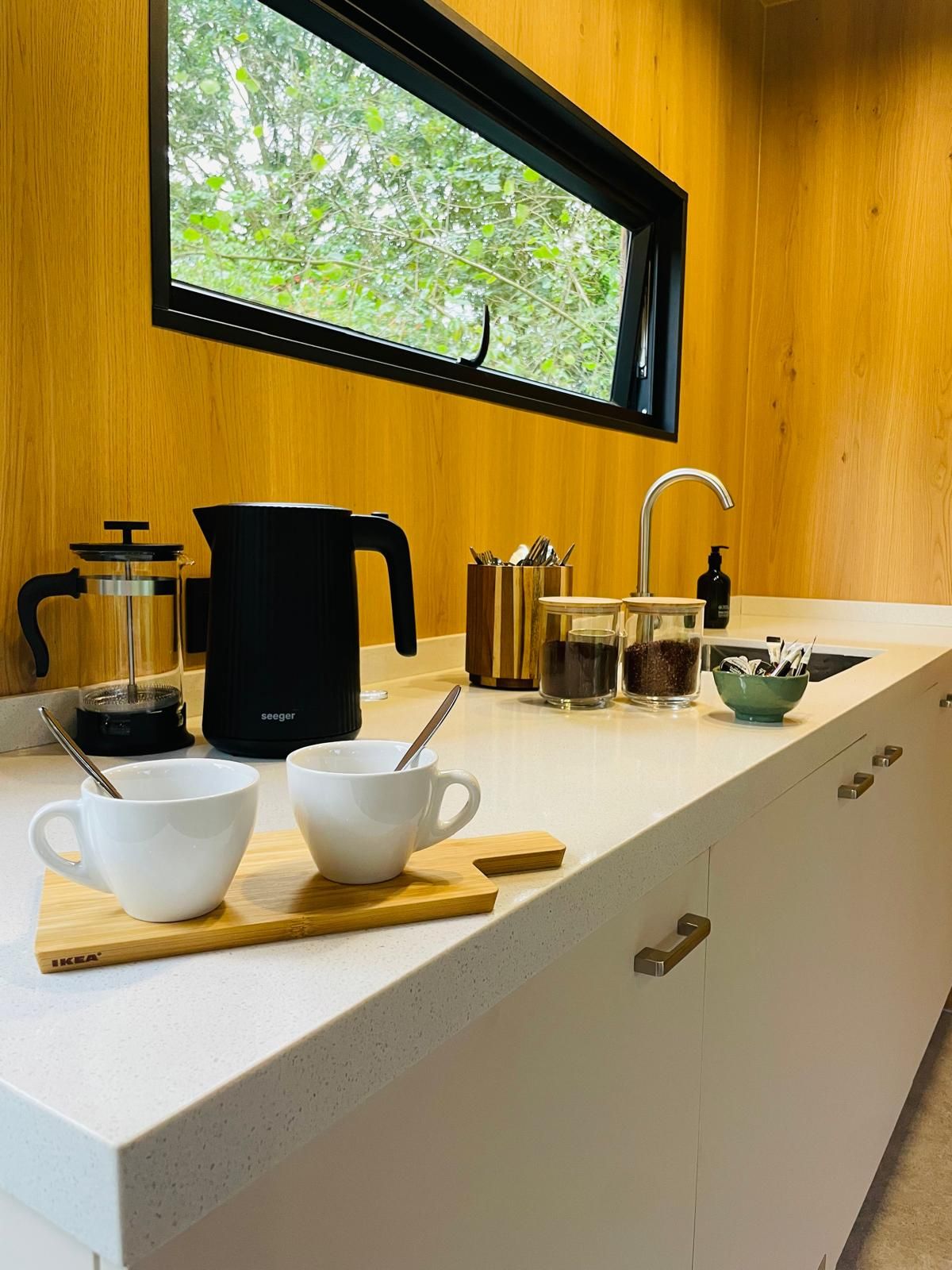 Cozinha com janela, cafeteira, chaleira e duas xícaras brancas sobre uma tábua de madeira.