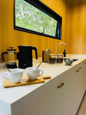 A kitchen with a window, a coffee maker, a kettle, and two white cups on a wooden board.