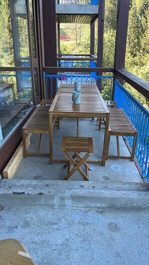Um deck de madeira ao ar livre com mesa e bancos, guarda-corpo azul e vista para árvores e um prédio ao fundo.
