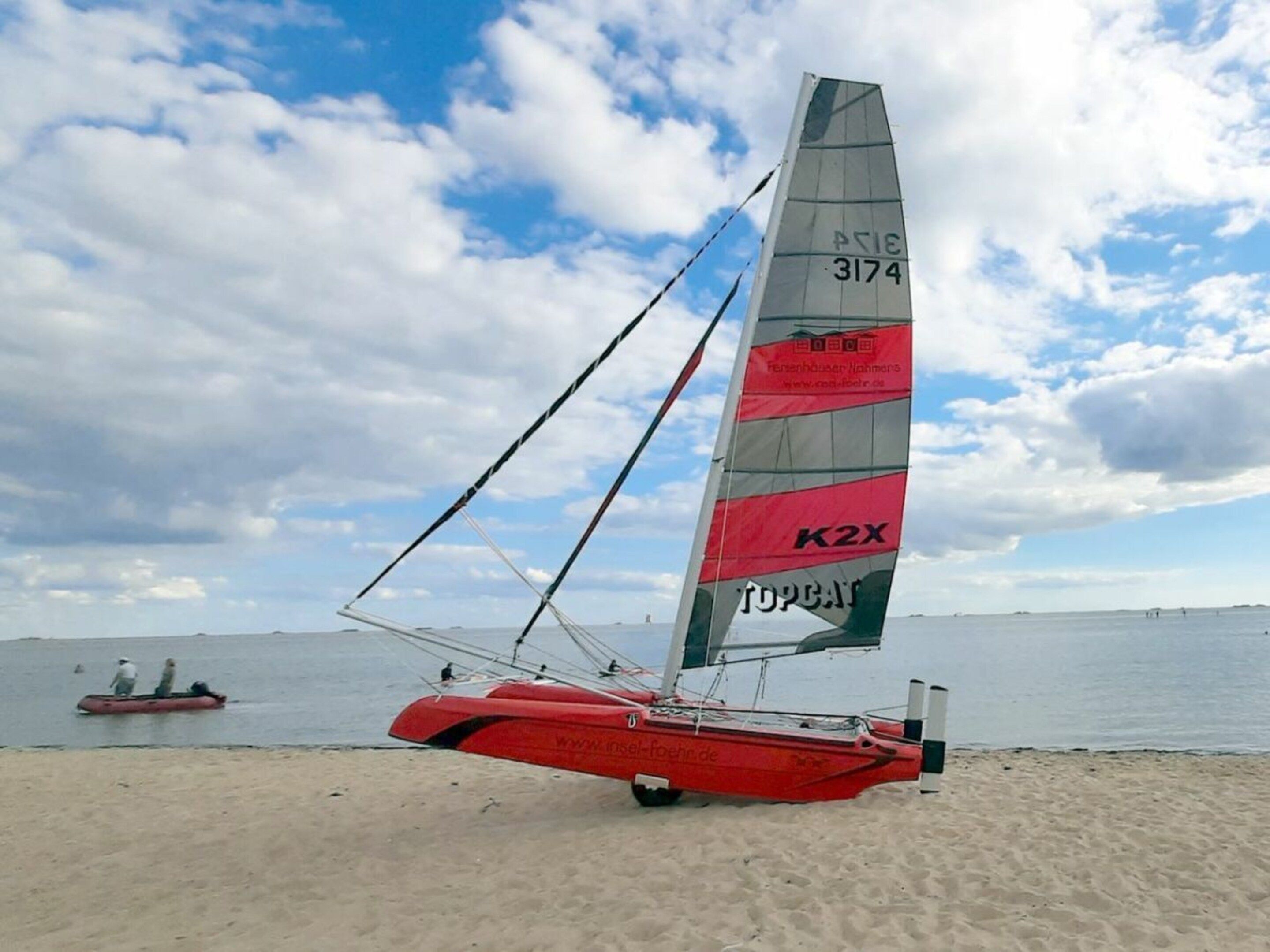 Een rode zeilboot met een hoge zeil aan een zandstrand, met kalm water en een bewolkte lucht op de achtergrond.