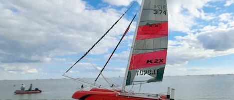 Een rode zeilboot met een hoge zeil aan een zandstrand, met kalm water en een bewolkte lucht op de achtergrond.