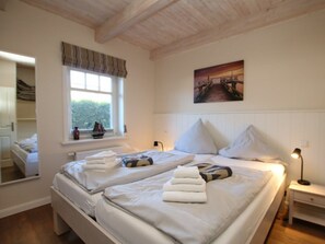 A double bed with pillows and folded towels, a bedside table with a lamp, a window with curtains, a painting on the wall, and a mirror reflecting part of the room. - Seeschwalbe at Susanne-fischer-weg 49 (Wyk auf Foehr)