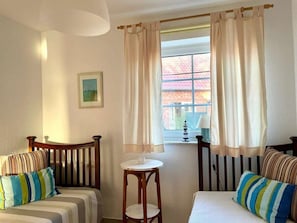 Ein Zimmer mit zwei Betten, einem kleinen Tisch, einer Lampe und einem Fenster mit Vorhängen.