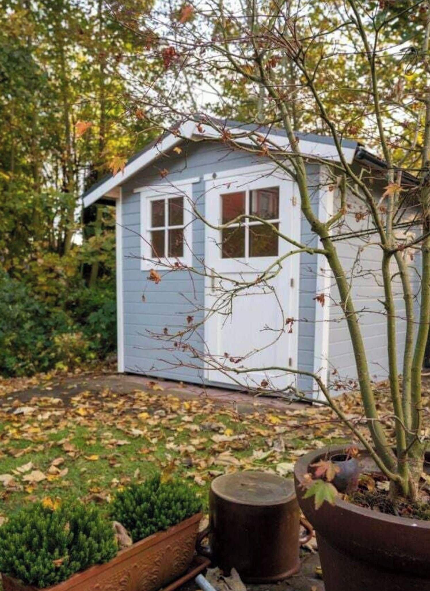 Un petit abri de jardin avec une porte et des fenêtres blanches, entouré d’arbres et d’un jardin où se trouvent une plante en pot et un baril en métal.