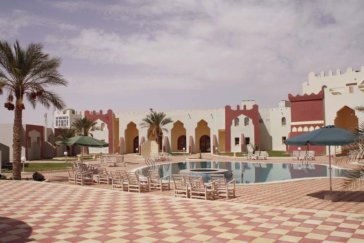 Uma área de piscina com espreguiçadeiras e guarda-sóis, rodeada por um edifício com galerias em arco e chão em mosaico.