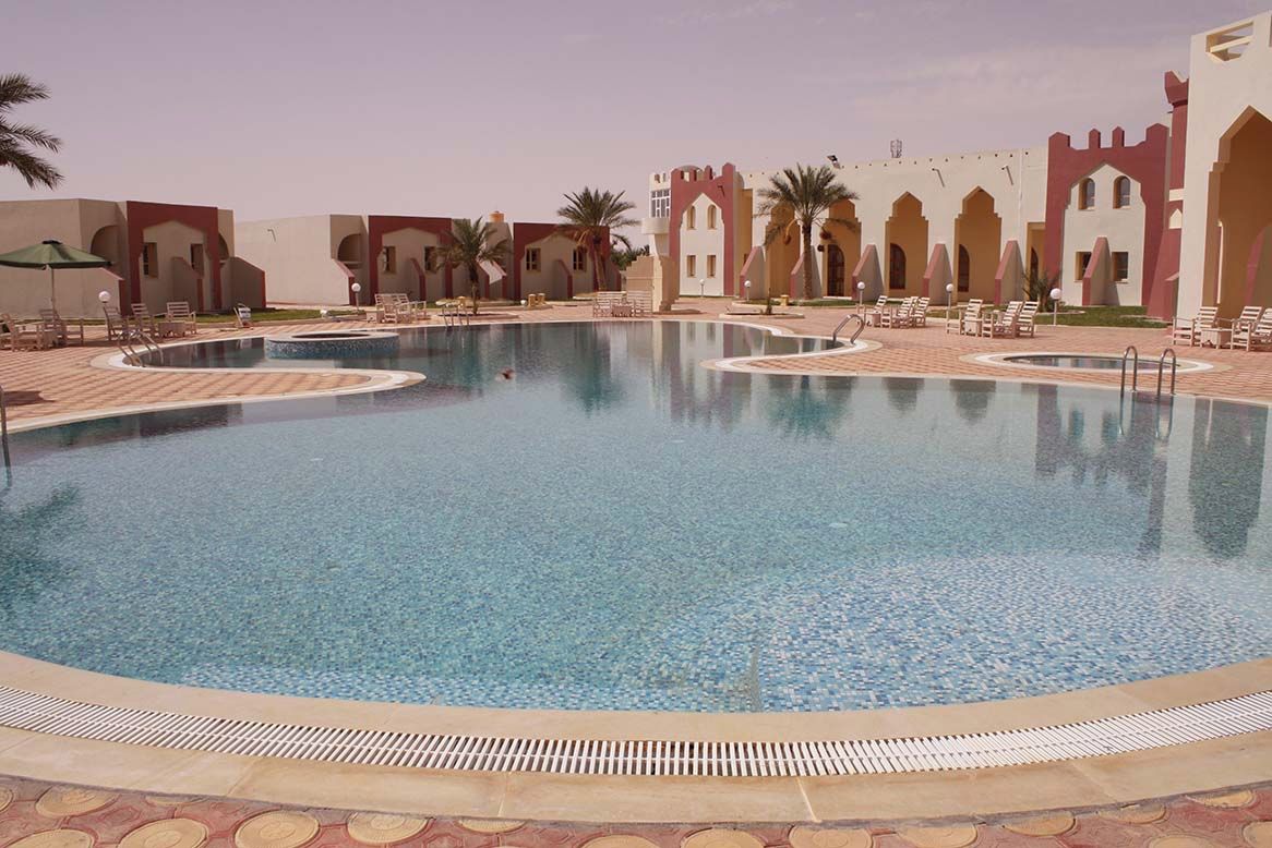 A swimming pool with a tiled deck, surrounded by lounge chairs and palm trees.