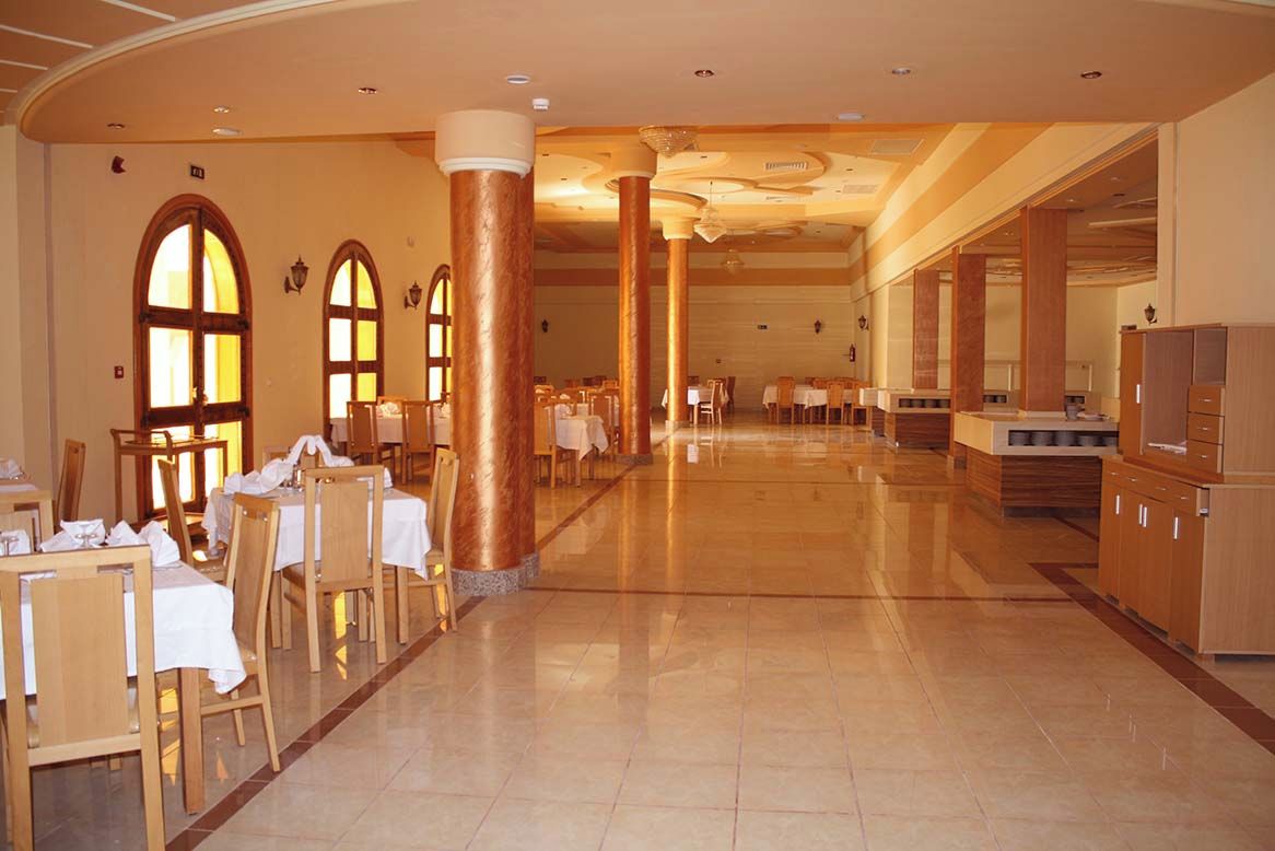 A spacious hotel dining area with multiple tables and chairs, large columns, and a modern kitchen area.