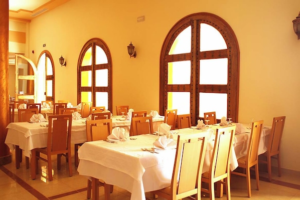 A dining area with multiple tables and chairs, large arched windows, and wall-mounted light fixtures.