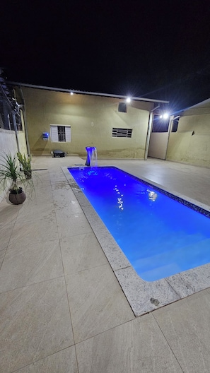 A rectangular swimming pool with a blue light feature, surrounded by a tiled patio area with a potted plant.