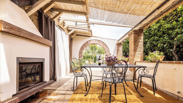 Eine Terrasse im Freien mit Tisch und Stühlen, einem Kamin und Blick auf einen Garten durch einen Bogen.