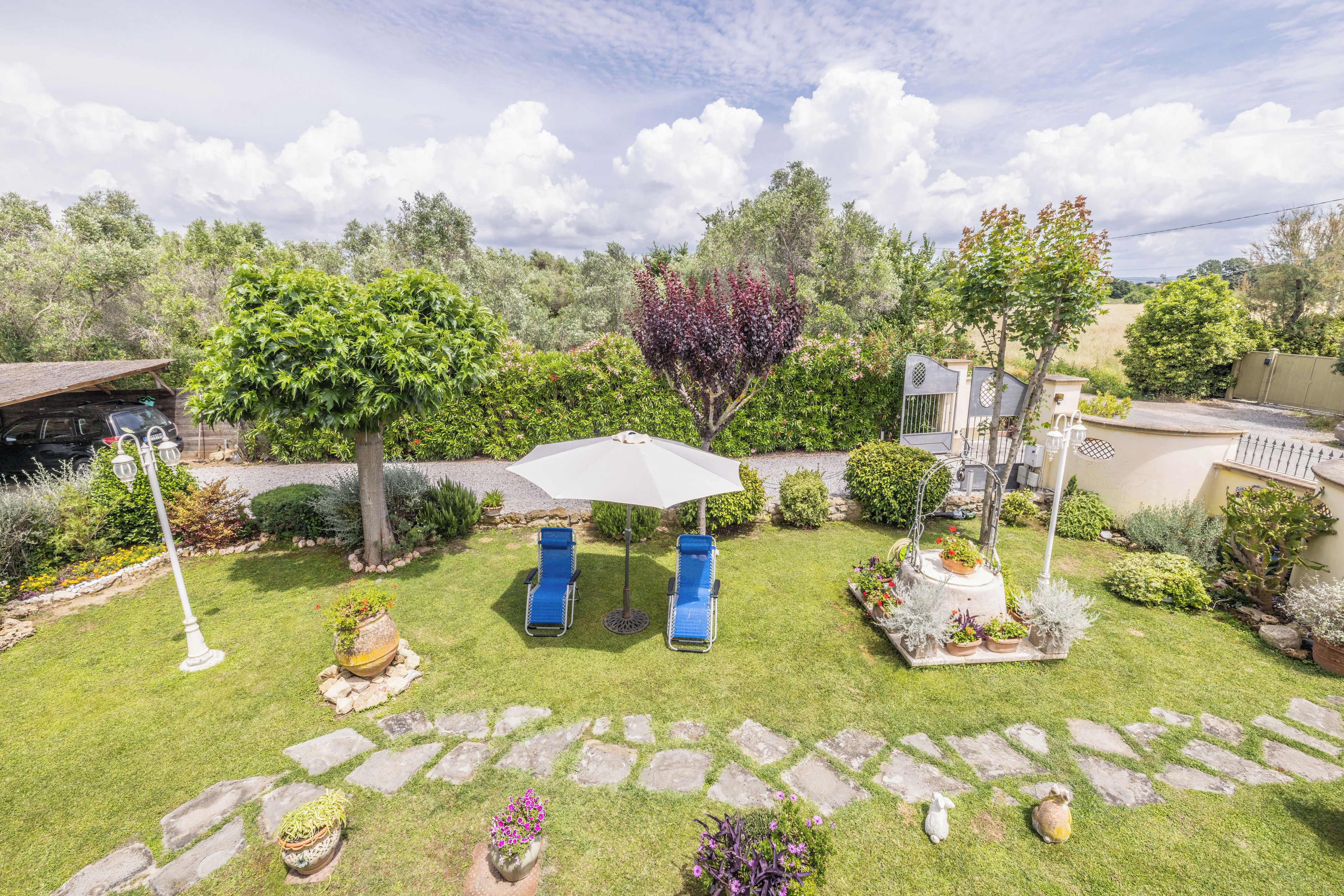 Un jardín bien cuidado con césped, dos sillones azules, una sombrilla blanca y diversas macetas con plantas.
