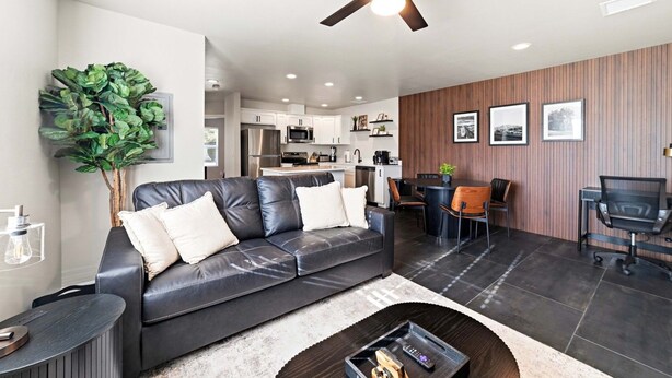 A modern living room with a leather sofa, dining table, and chairs, a ceiling fan, and a kitchen in the background. - Work-Ready Stay with Desk + ALLO Fiber WiFi (Kingman)