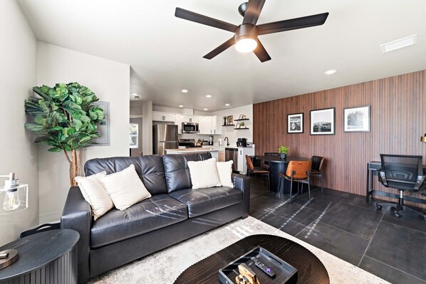 A modern living room with a leather sofa, dining table, and chairs, a ceiling fan, and a kitchen in the background. - Work-Ready Stay with Desk + ALLO Fiber WiFi (Kingman)