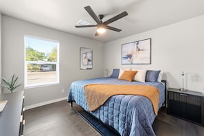 A bedroom with a bed, ceiling fan, artwork, and a window with a view.