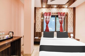 Hotellihuone, jossa on sänky, yöpöytä, jossa on vedenkeitin, toinen yöpöytä ja ikkuna, jossa on verhot.