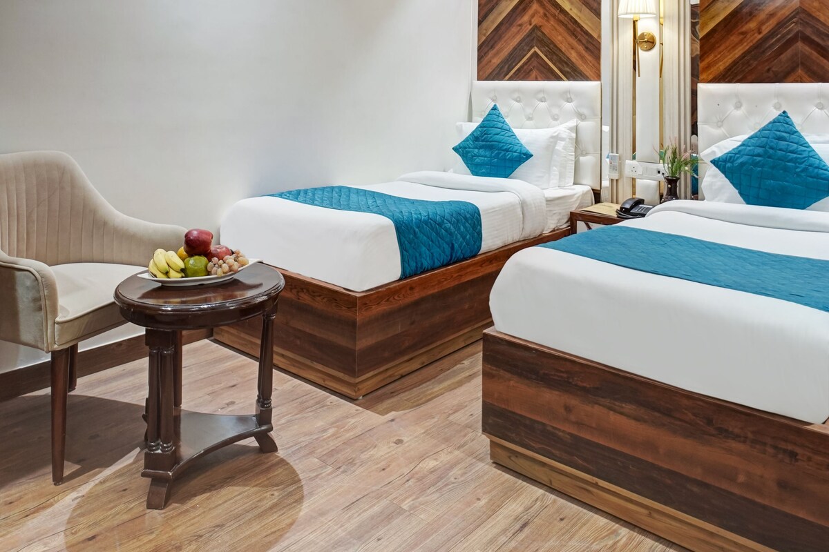A hotel room with two beds, a small table with fruit, a chair, and a decorative wall panel.