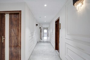 A hallway with white walls, wooden doors, and a marble floor. - Hotel Saina International - Newly Built  (New Delhi)