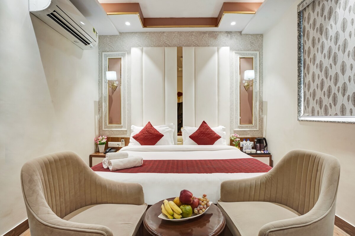 Une chambre d’hôtel comprenant un lit, deux fauteuils, une table de chevet avec un panier de fruits et un miroir orné d’un cadre décoratif.