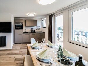 Eine moderne Küche mit integrierten Geräten, eine Essecke mit einem Holztisch für vier Personen und ein großes Fenster mit Blick ins Freie.