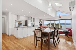 Una cocina moderna con muebles blancos, un comedor con mesa y sillas de madera, y vistas al exterior a través de amplios ventanales.