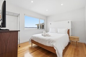 Un dormitorio con una cama, una mesita de noche de madera, un televisor sobre un soporte, una ventana con vistas y una lámpara en el techo.