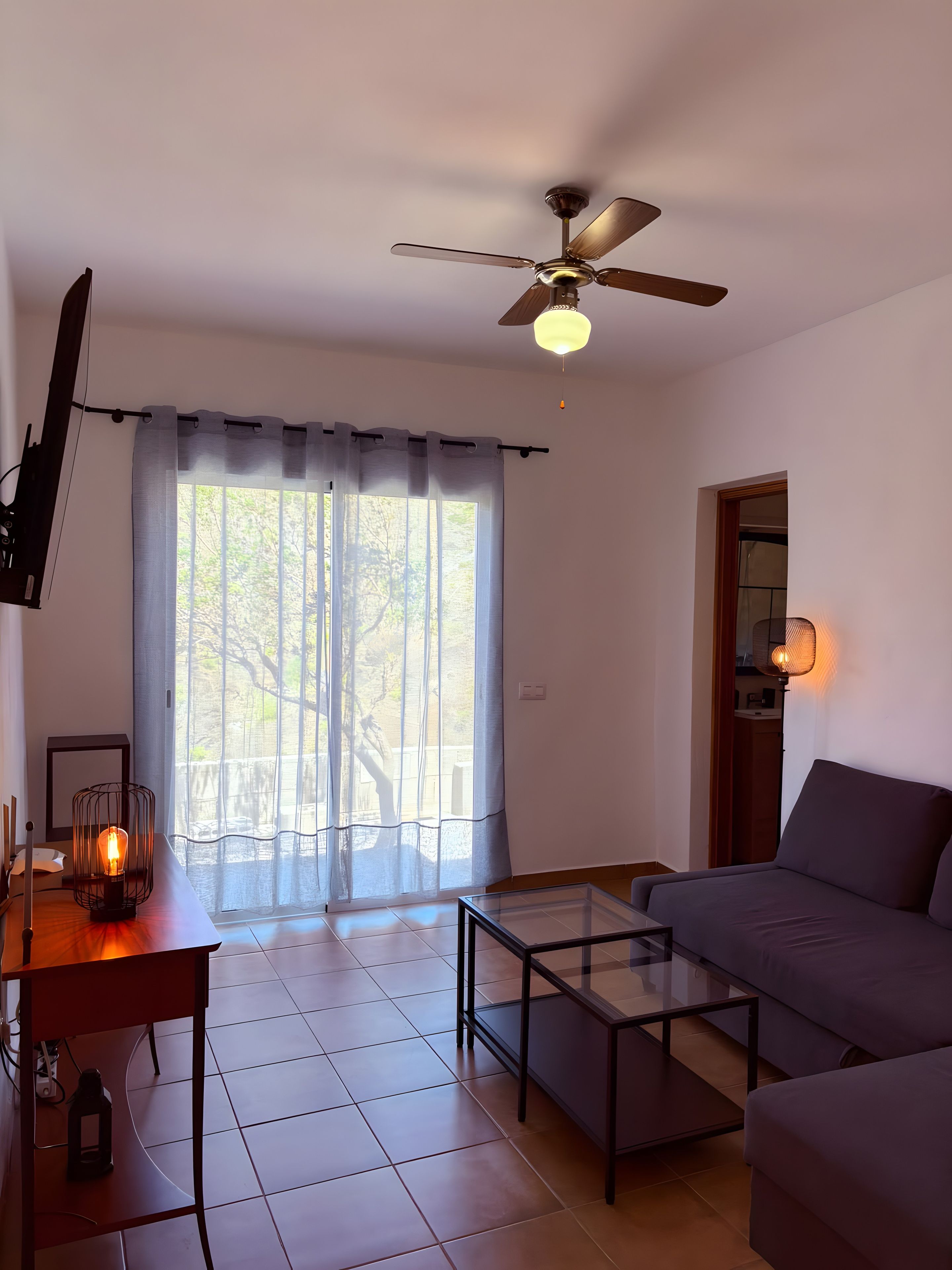 Un salon doté d’un ventilateur de plafond, d’un canapé, d’une table basse en verre et d’un téléviseur fixé au mur.