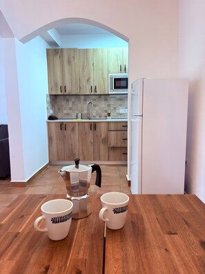 Un coin repas avec une table en bois, deux tasses blanches ornées de motifs noirs, une cafetière et, au fond, une cuisine.