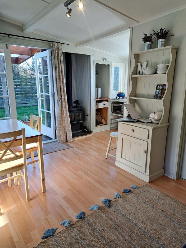 A room with a wooden dining table, a white cabinet with shelves, and a view of the outdoors through large windows.