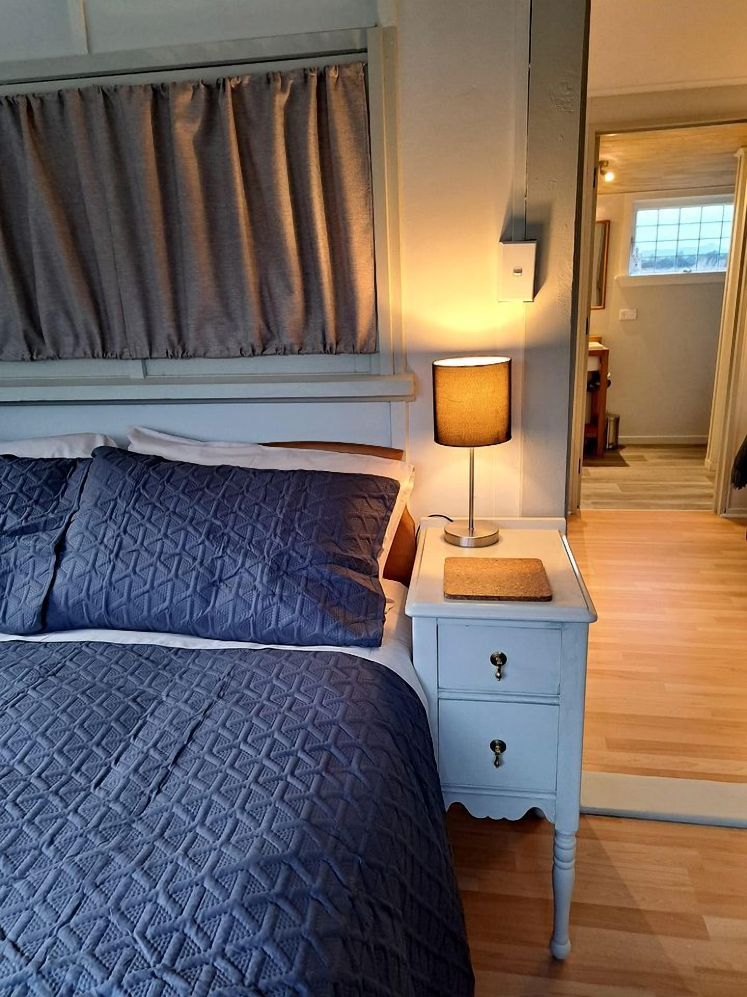 A bedroom with a bed, a nightstand with a lamp, and a door leading to another room.