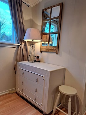 A room with a dresser, a lamp, a stool, a window with curtains, and a mirror.