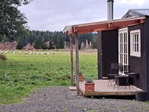 A small cabin with a porch, a chimney, and a view of a grassy field with sheep.
