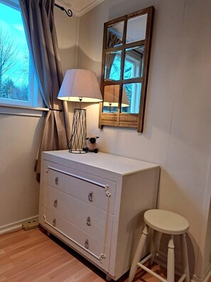 A room with a dresser, a lamp, a stool, a window with curtains, and a mirror.