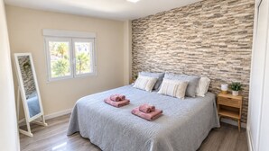 Un dormitorio con pared de piedra, una cama con toallas enrolladas, una mesita de noche y una ventana con vista a palmeras.