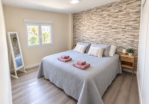 A bedroom with a stone wall, a bed with rolled towels, a bedside table, and a window with a view of palm trees. - Vrbo Property (Costa Adeje)