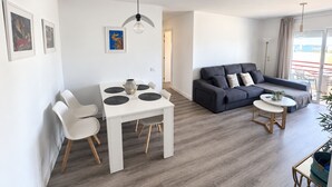 Una sala de estar moderna con un sofá gris, una mesa de comedor blanca con cuatro sillas y una mesa redonda pequeña con una planta.