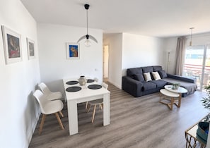 A modern living room with a grey sofa, a white dining table with four chairs, and a small round table with a plant. - Vrbo Property (Costa Adeje)