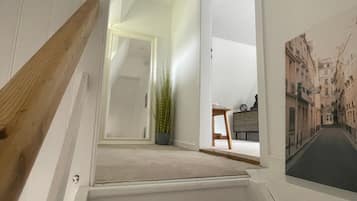 Una escalera con barandilla de madera que lleva a una plataforma. Hay una planta en maceta sobre la plataforma y un cuadro enmarcado en la pared.
