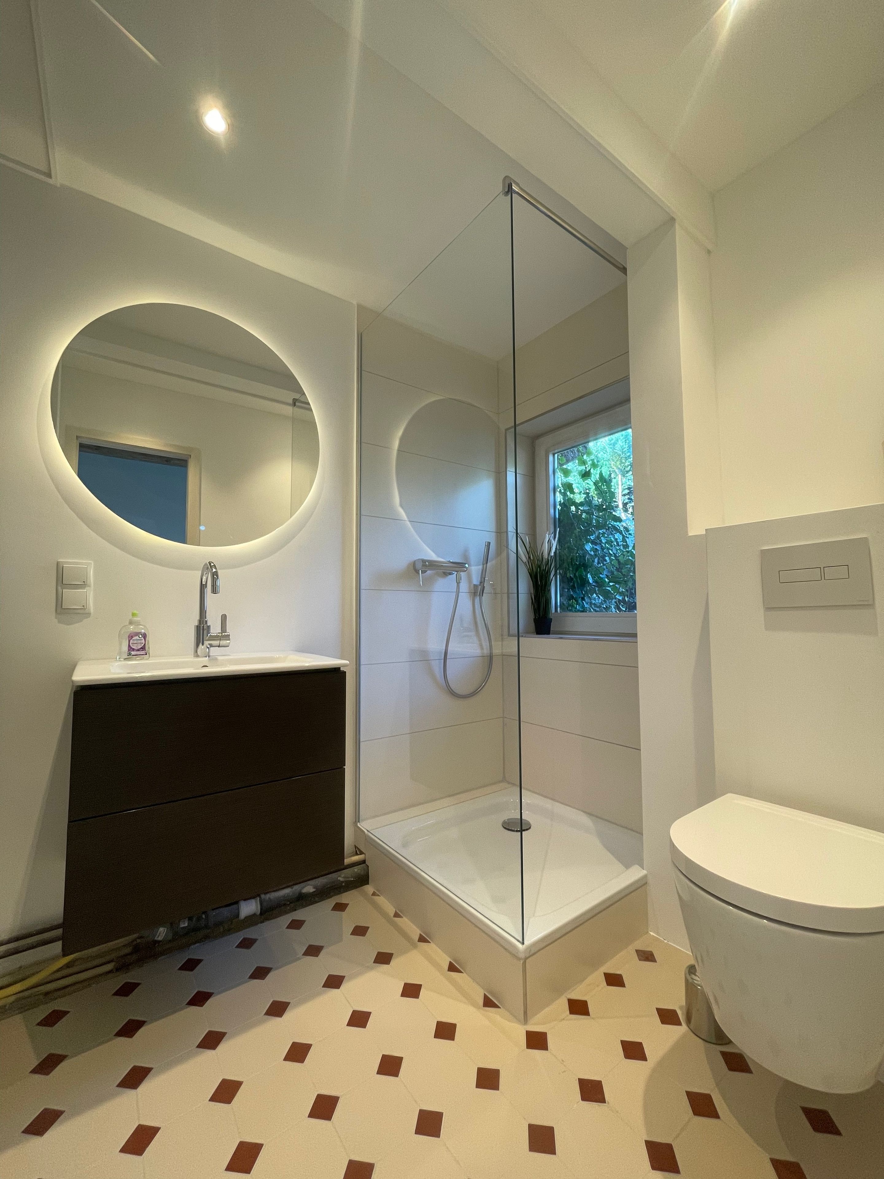 Ein modernes Badezimmer mit einer gläsernen Duschkabine, einem runden Spiegel, einer weißen Toilette und einem gemusterten Boden.