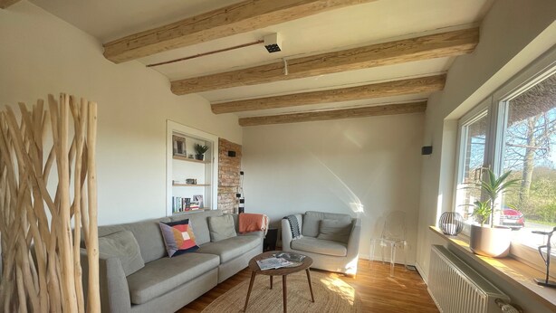 Ein Wohnzimmer mit grauem Sofa, einem runden Couchtisch, Holzbalken an der Decke, einer Heizung und einem Fenster mit Blick nach draußen.