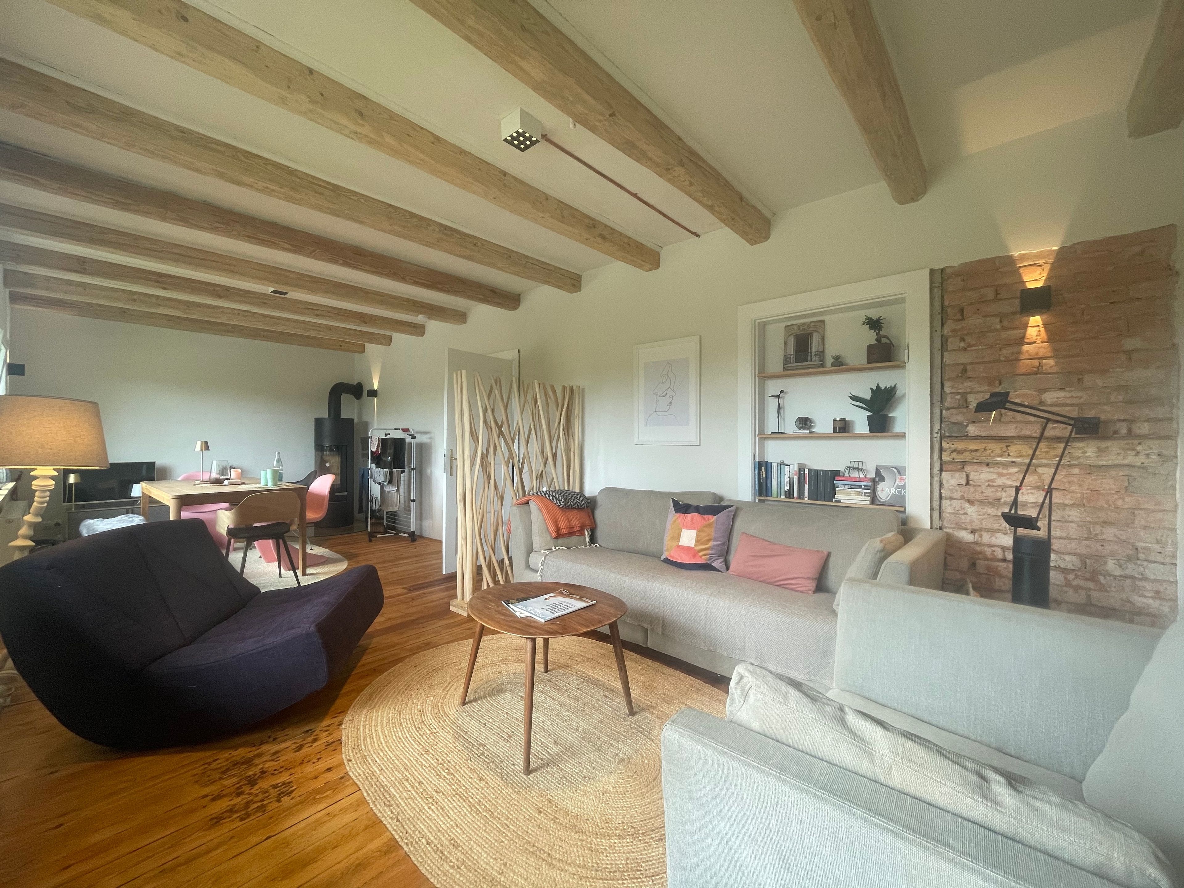 Ein Wohnzimmer mit Sofa, Sessel und Couchtisch. Ein Kamin, eine Bücherwand und eine Lampe sind ebenfalls vorhanden.