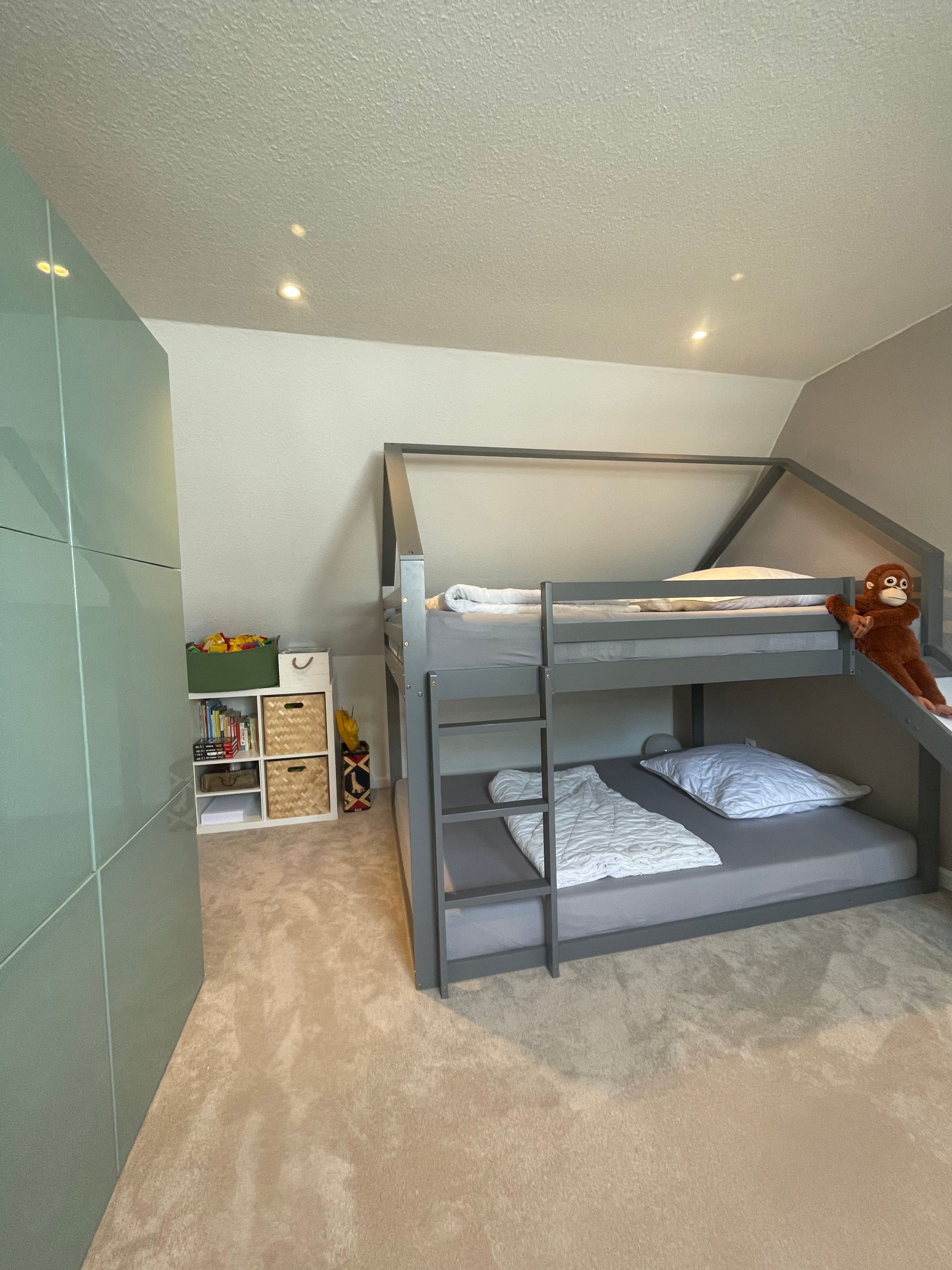 Ein Etagenbett mit Stauraum darunter, ein Regal mit Büchern und Spielzeug sowie ein kleiner Tisch mit Lampe.