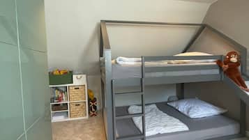 Una cama litera con un espacio de almacenamiento debajo, un estante con libros y juguetes, y una mesita con una lámpara.