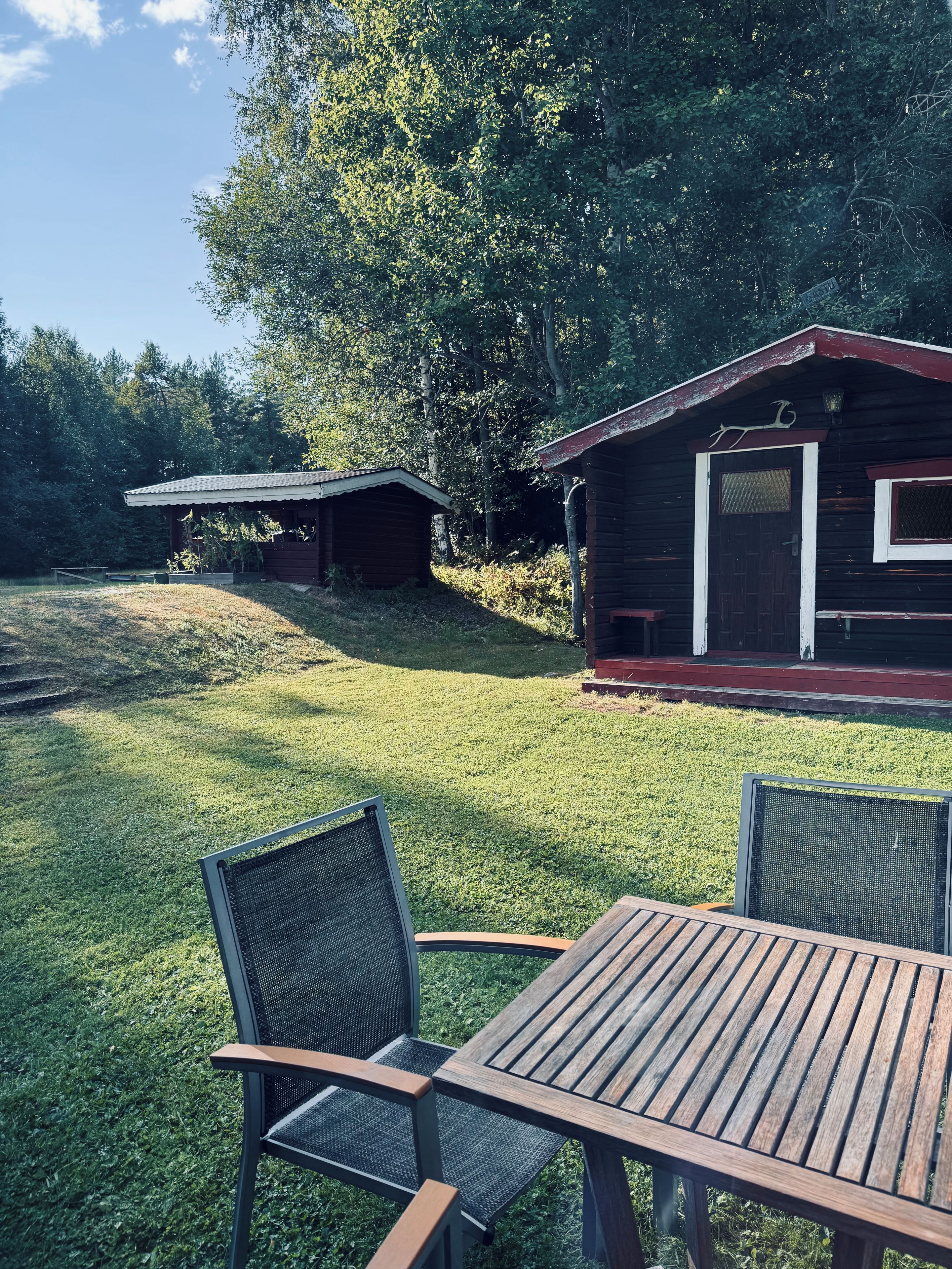 En träveranda med två stolar och ett bord, omgiven av en gräsyta med en liten stuga i bakgrunden.