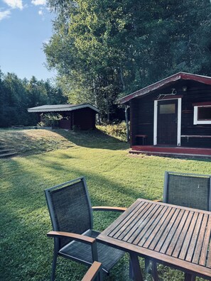 A wooden deck with two chairs and a table, surrounded by a grassy area with a small cabin in the background. - Vrbo Property (Tärnsjö)