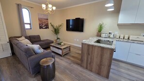 A modern living room with a sofa, coffee table, and a small round stool. A flat-screen TV is mounted on the wall, and there is a kitchen area with a countertop and cabinets.
